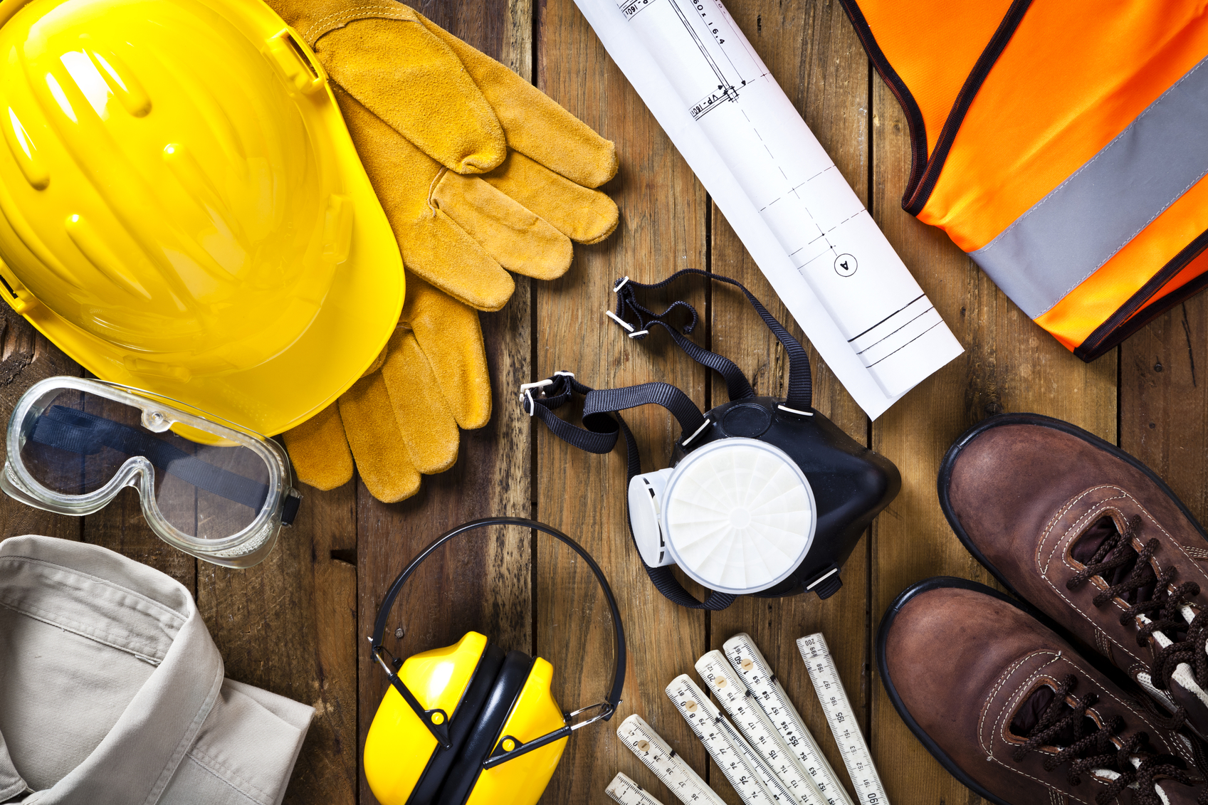 Personal protective workwear and blueprint shot directly from above on rustic wood background. The protective workwear includes hard hat, gloves, earmuff, goggles, steel toe shoes, and safety vest. Predominant colors: yellow and brown. DSRL studio photo taken with Canon EOS 5D Mk II and EF 100mm f/2.8L Macro IS USM